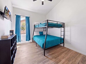 Ein Etagenbett mit blauer Matratze, Holzboden, Deckenventilator und Fenster mit Vorhängen.