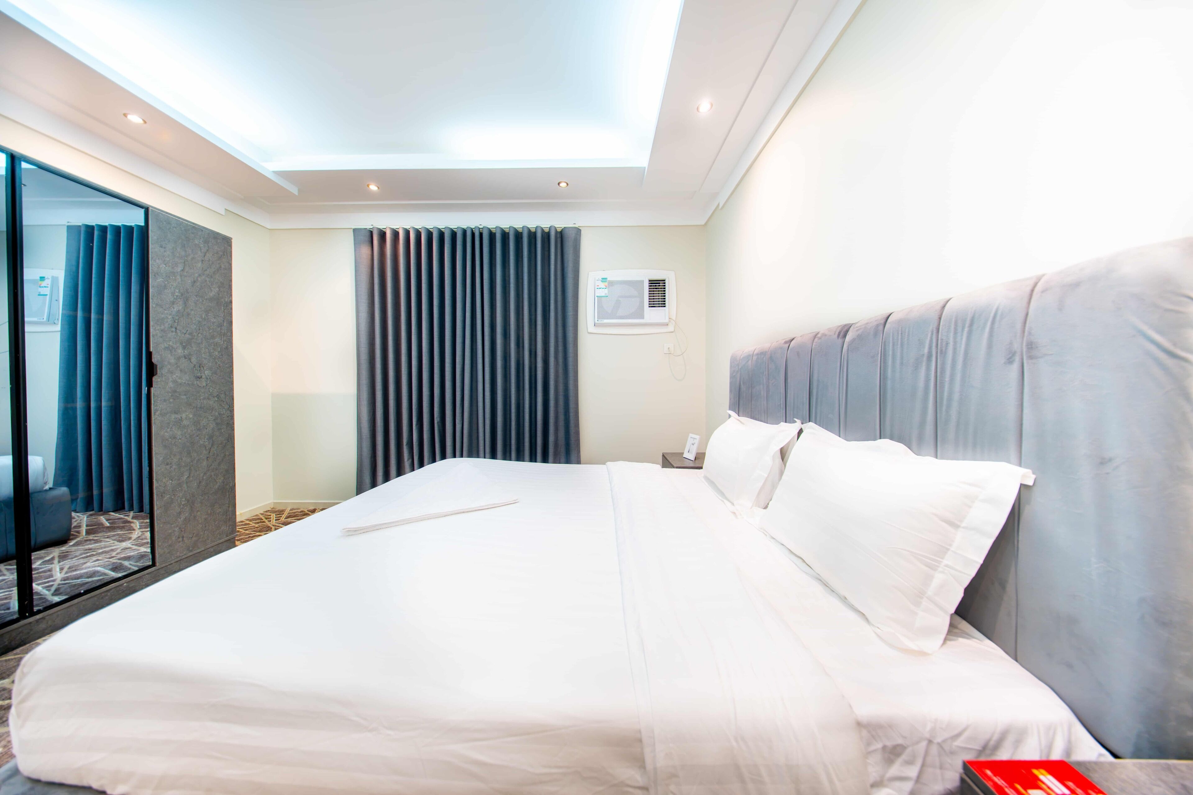 A hotel room with a large bed, a glass partition, a window with curtains, and an air conditioning unit.