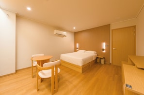 A hotel room with a bed, a wooden desk, a chair, a small table, and a wall-mounted air conditioning unit.