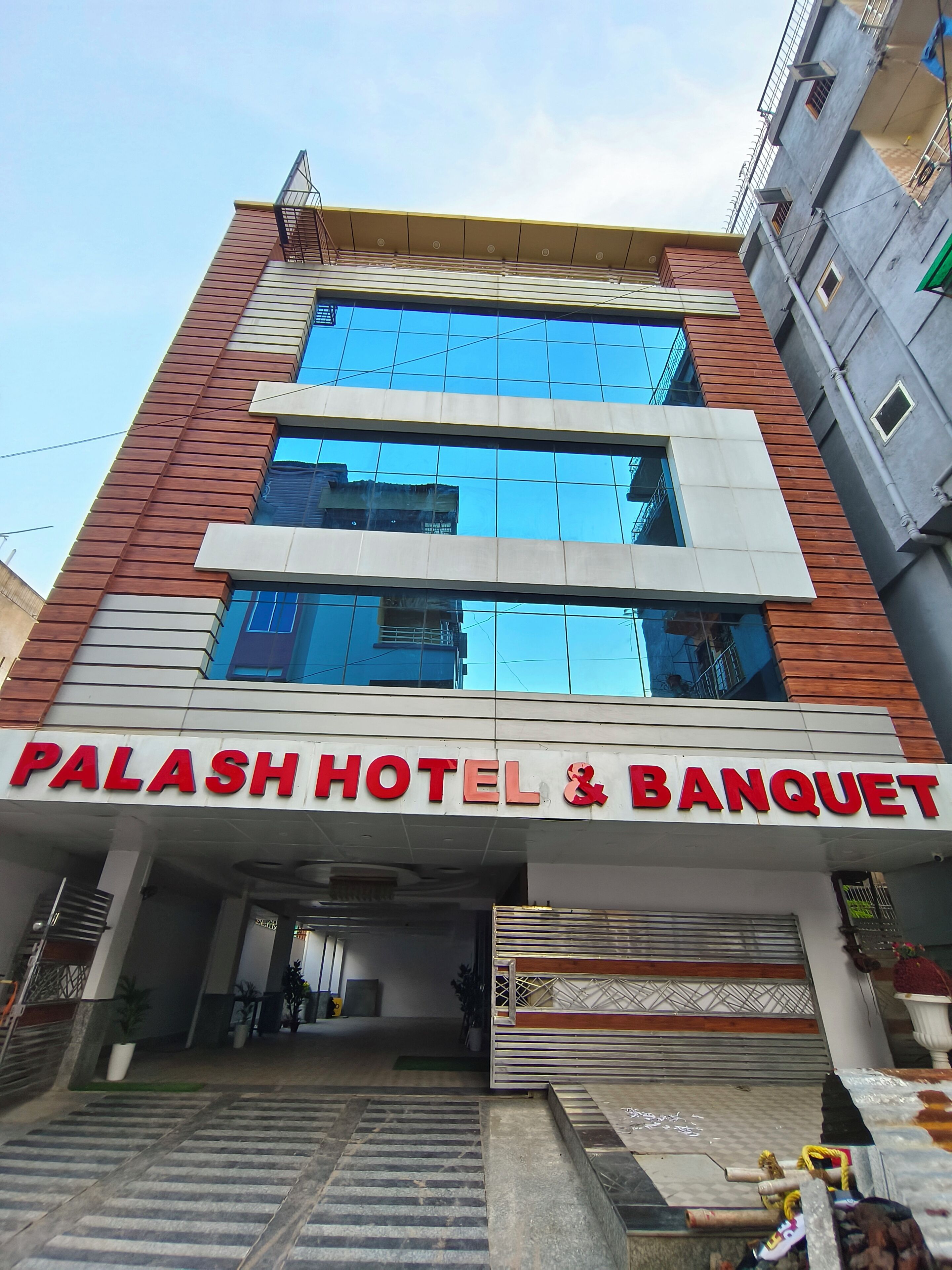 Um hotel com fachada moderna, grandes janelas e uma placa destacada com a inscrição 'PALASH HOTEL & BANQUET'.