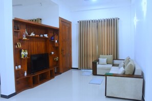 A living room with a flat-screen TV, wooden entertainment unit, beige sofa, and a large window with curtains.