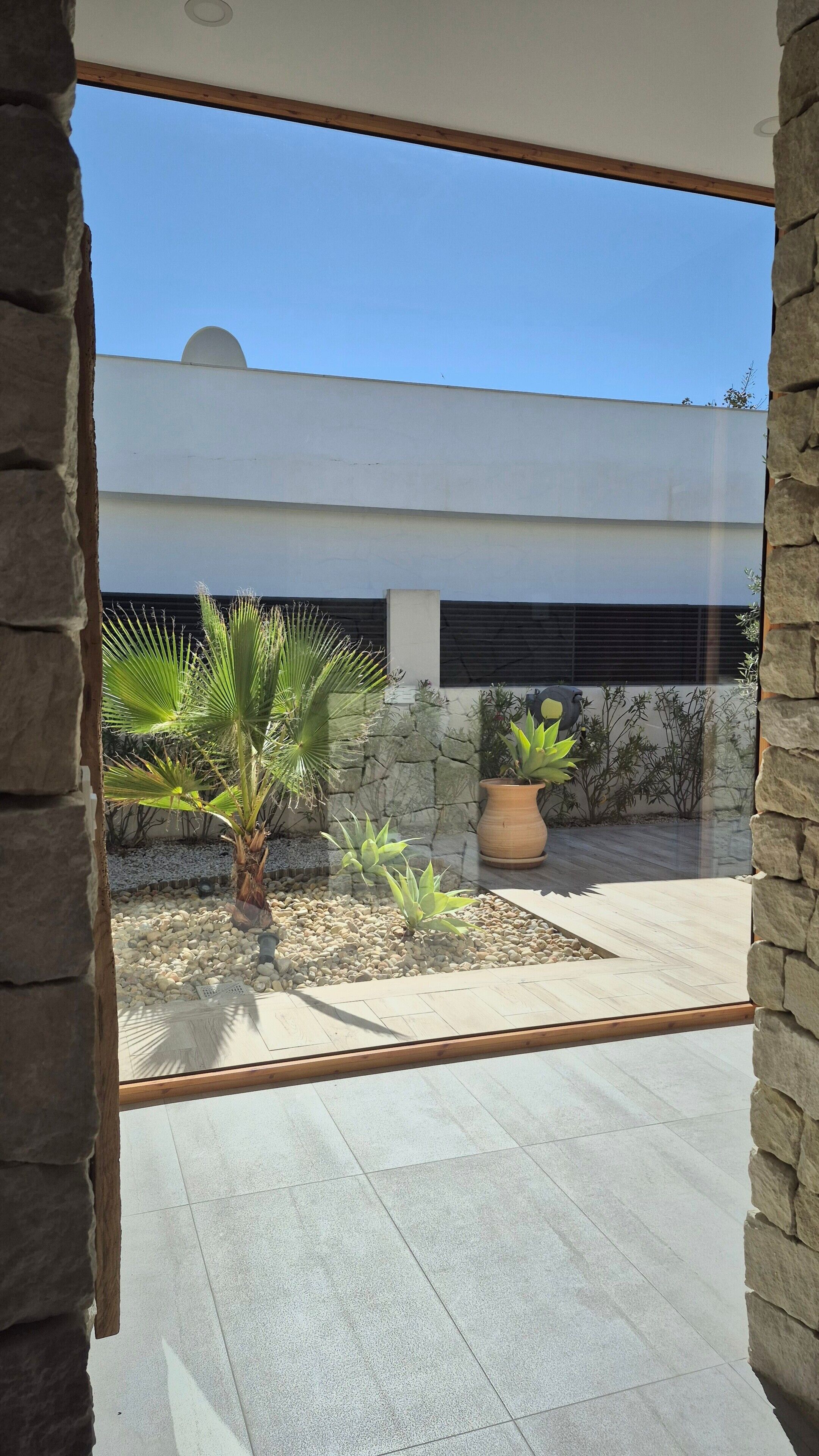 Un bâtiment moderne à la façade en pierre, sous un ciel d’un bleu éclatant, et un jardin agrémenté de diverses plantes et d’un grand pot.