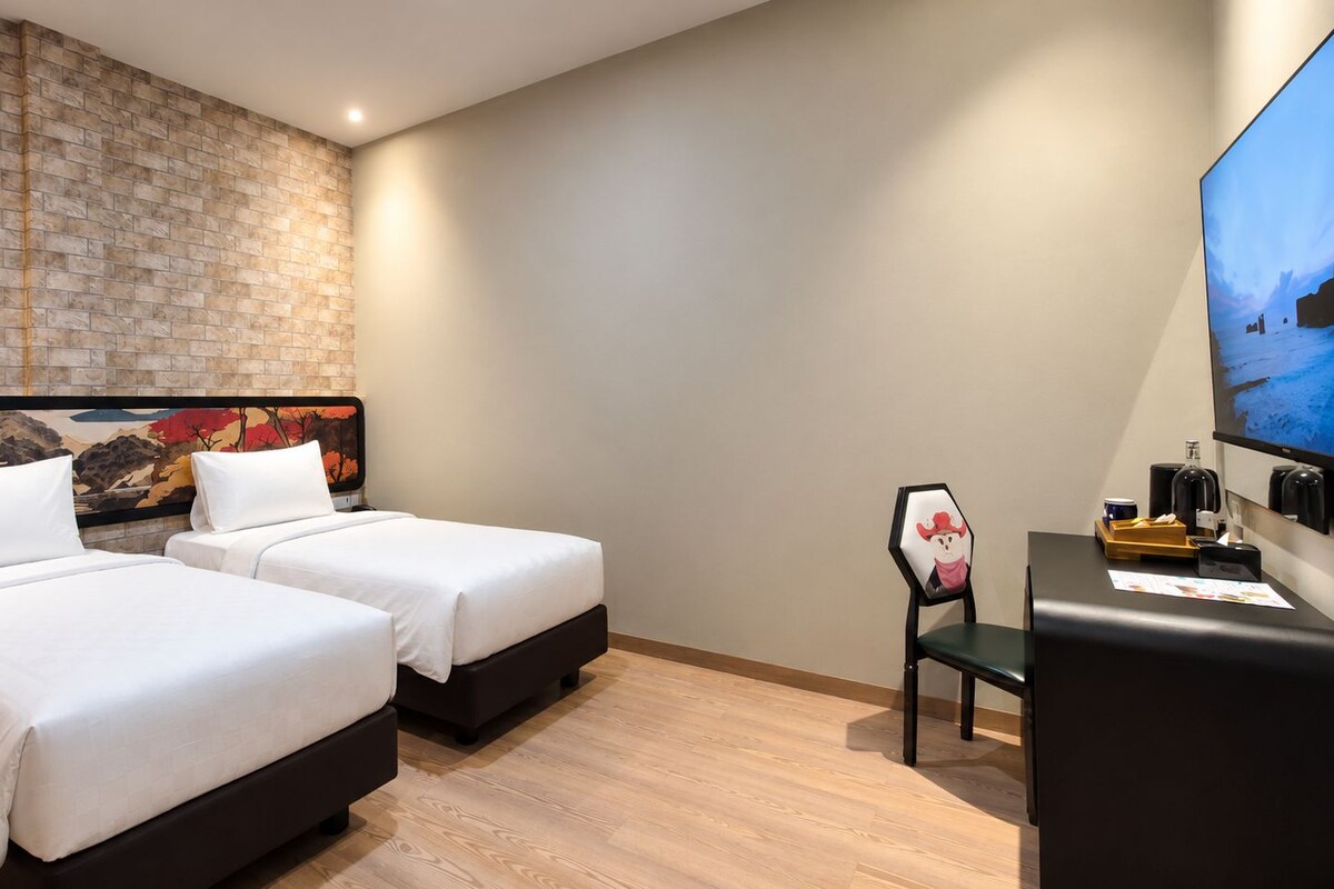 Habitación de hotel con dos camas, pared de ladrillo decorativa, televisor de pantalla plana y escritorio con silla.