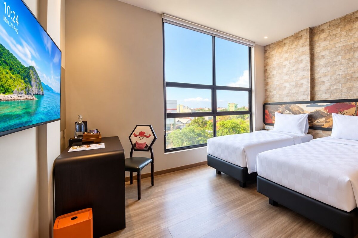 Habitación de hotel con dos camas, un escritorio con silla, un televisor y un ventanal con vistas a la ciudad.