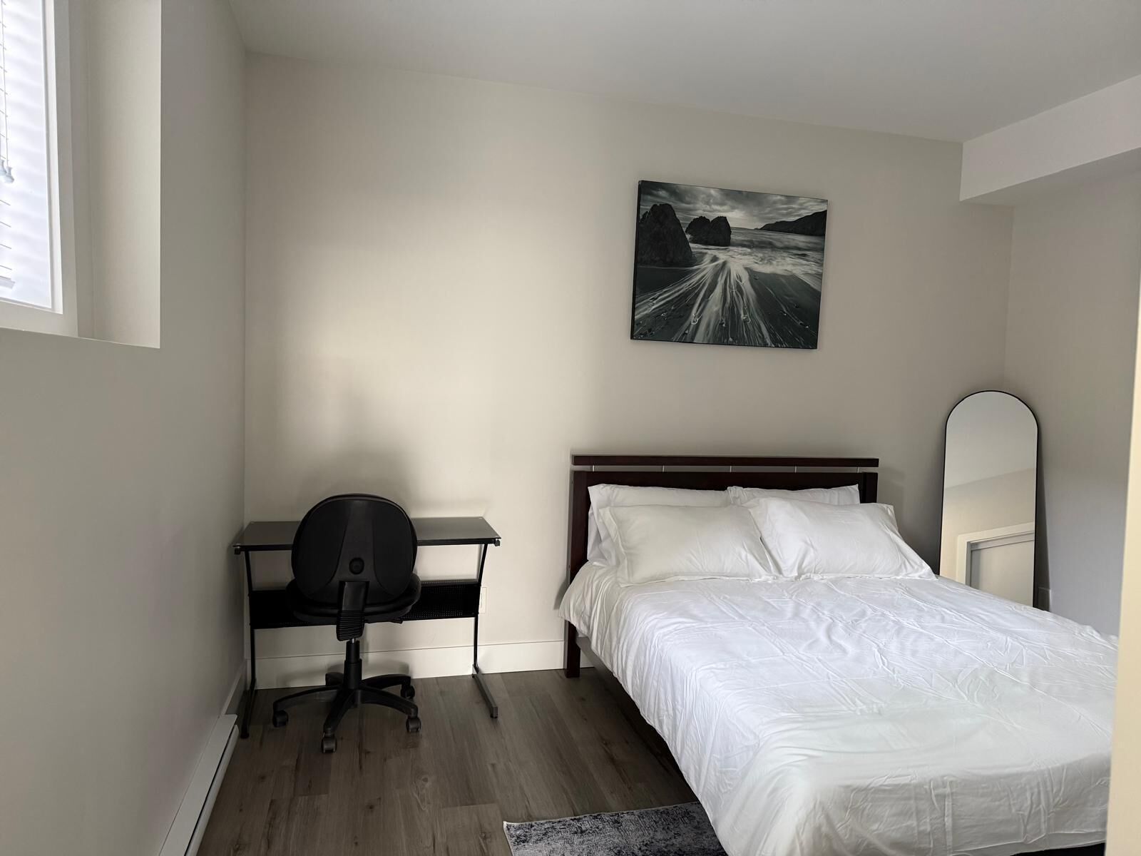 A bedroom with a bed, a desk, a chair, a painting, and a mirror.