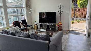 Un salon moderne avec un canapé gris, un fauteuil noir, un téléviseur à écran plat, un vase décoratif avec des fleurs et une vue sur l’extérieur.