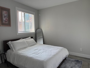 A bedroom with a bed, a window with blinds, a wall-mounted mirror, and a framed picture.