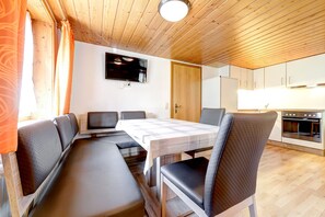 Ein Essbereich mit Tisch und Stühlen, einem an der Wand befestigten Fernseher sowie einer Küche mit Schränken und einem Herd.