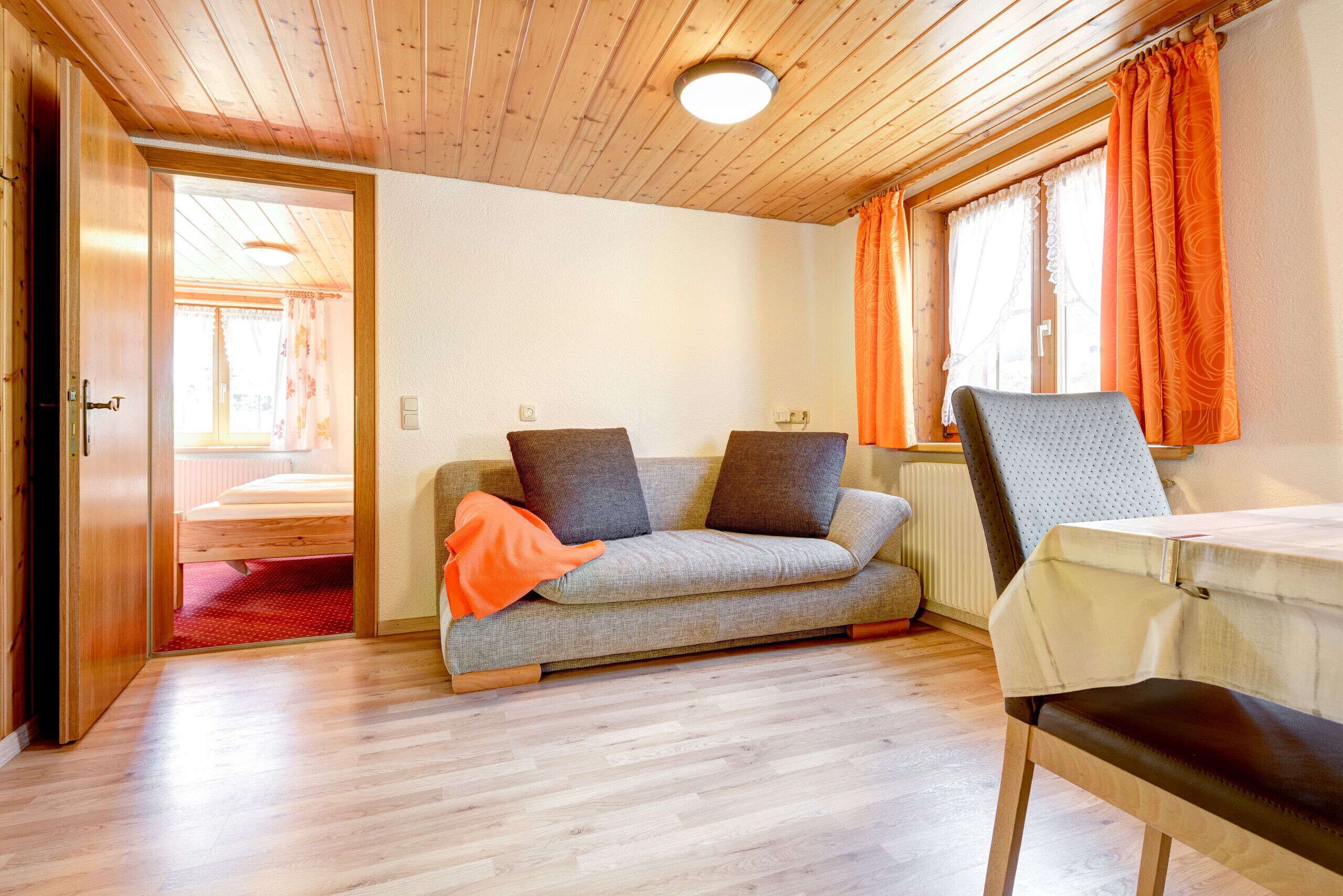 A room with a wooden ceiling, a sofa with cushions, a chair, a table, and a window with curtains.