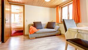 A room with a wooden ceiling, a sofa with cushions, a chair, a table, and a window with curtains. - Ferienwohnung Brügga (Brand)