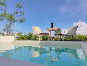 Ein Poolbereich mit Liegestühlen und einem Tisch, umgeben von Grünpflanzen und blauem Himmel.