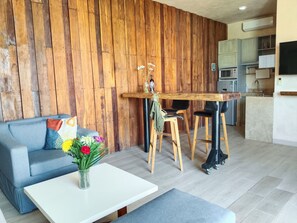 A modern living space with a wooden accent wall, a grey sofa, a white coffee table, and a kitchen area with a microwave, sink, and cabinets. - Dpto 1rec PH polo 15 (Playa del Carmen)