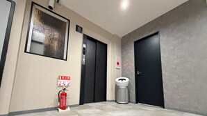 Corredor com extintor, lixeira e porta com luz de emergência vermelha.