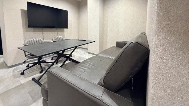 Uma sala de conferências com uma mesa grande, várias cadeiras, um sofá e uma TV de tela plana fixada na parede.