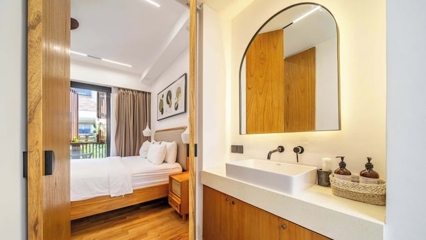 Un dormitorio moderno con piso de madera, una cama con ropa blanca, un espejo grande y un baño con lavamanos blanco y muebles de madera.