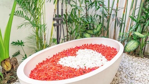Un área de spa al aire libre con un amplio jacuzzi ovalado lleno de piedras y pétalos de flores, rodeado de exuberante follaje tropical.