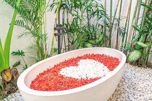 Ein Außen-Wellnessbereich mit einem großen, ovalen Whirlpool, gefüllt mit Kieselsteinen und Blütenblättern, umgeben von üppiger tropischer Vegetation.