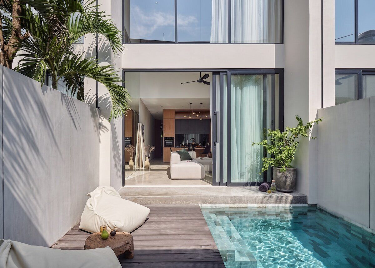 A modern outdoor pool area with a wooden deck, seating, and a view into the house.