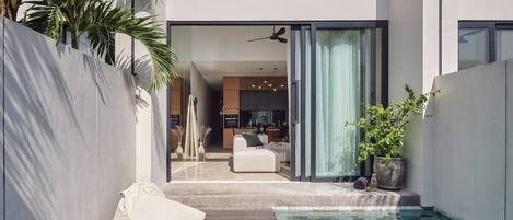 A modern outdoor pool area with a wooden deck, seating, and a view into the house.