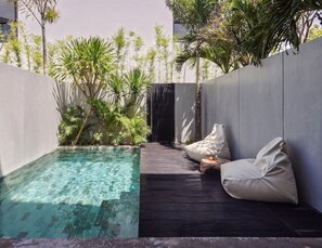 Un espace piscine extérieur moderne, doté d’une terrasse en bois sombre, d’un coin salon blanc avec des coussins moelleux et d’une végétation tropicale luxuriante.