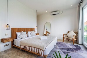 A bedroom with a bed, bedside tables, a chair, a mirror, and an air conditioning unit. - Tropical 3BR Canggu Stay w Pool, Balcony & Office (Kuta Utara)