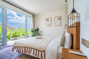 A bedroom with a bed, a bedside table, a lamp, and a view of greenery outside. - Tropical 3BR Canggu Stay w Pool, Balcony & Office (Kuta Utara)