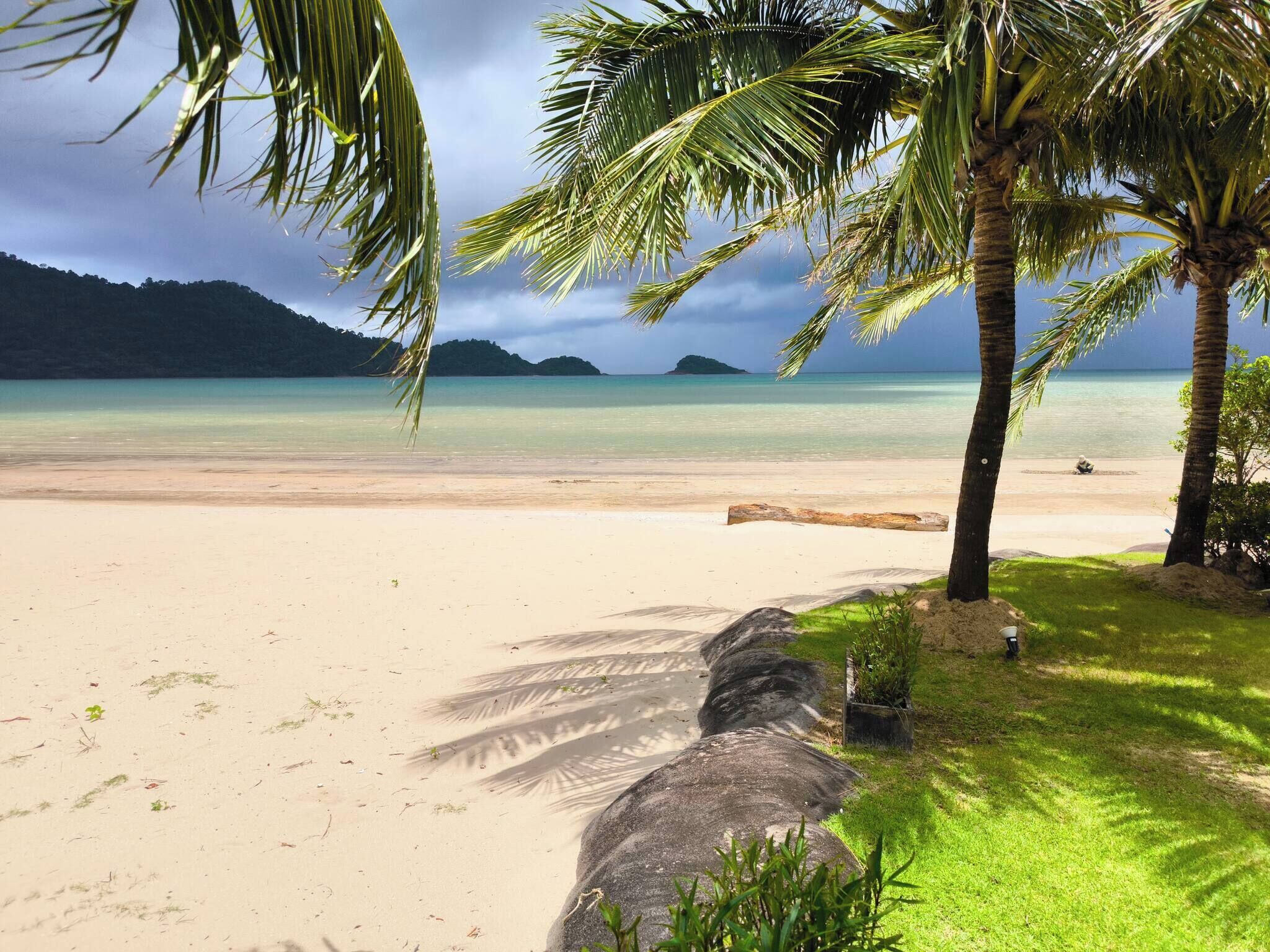 A beach with clear turquoise water, palm trees, and a large rock formation.