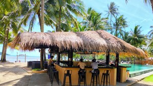 A beachside bar with a thatched roof, outdoor seating, and a view of the ocean.