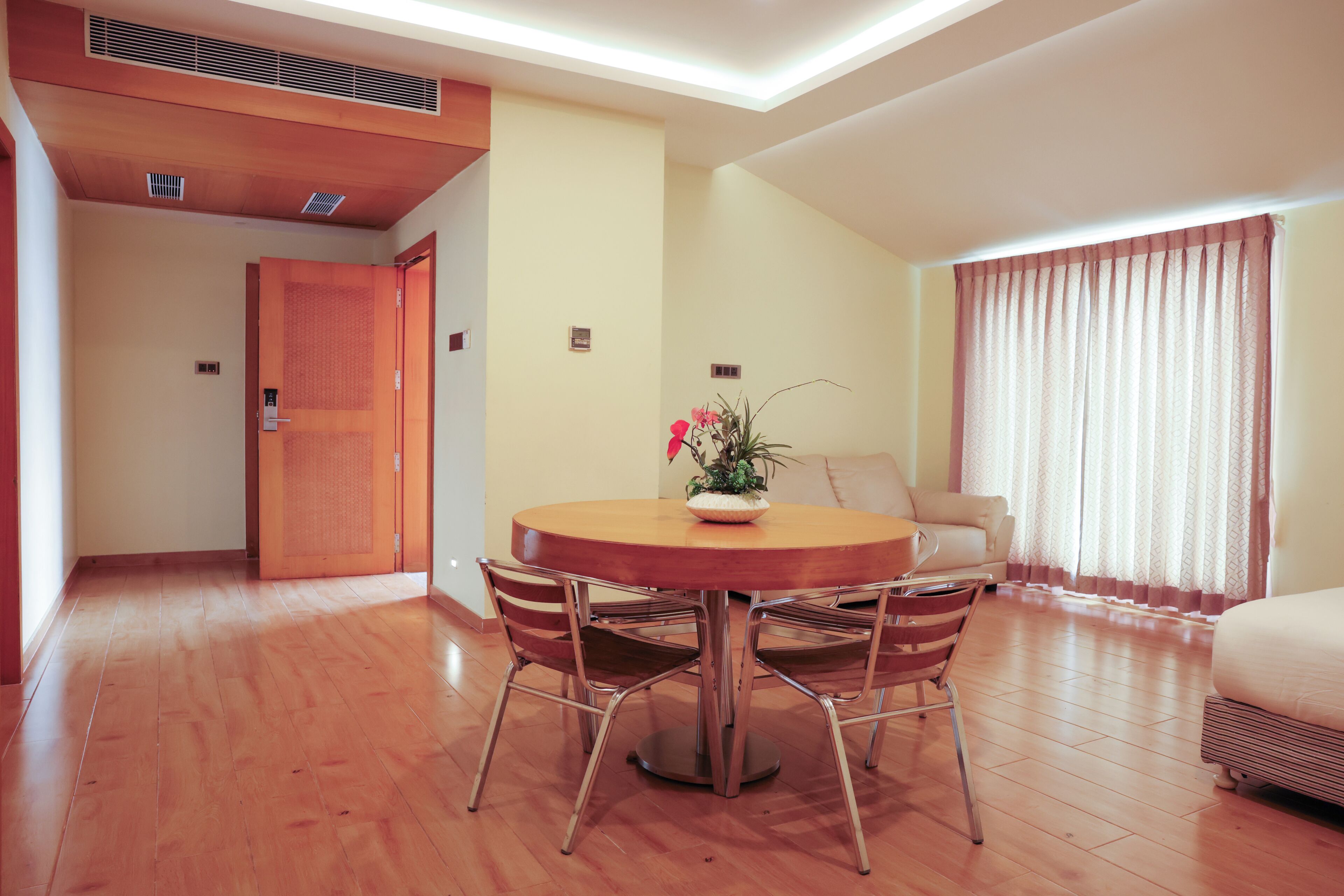 A room with a wooden floor, a round dining table with chairs, a potted plant, a sofa, and a window with curtains.