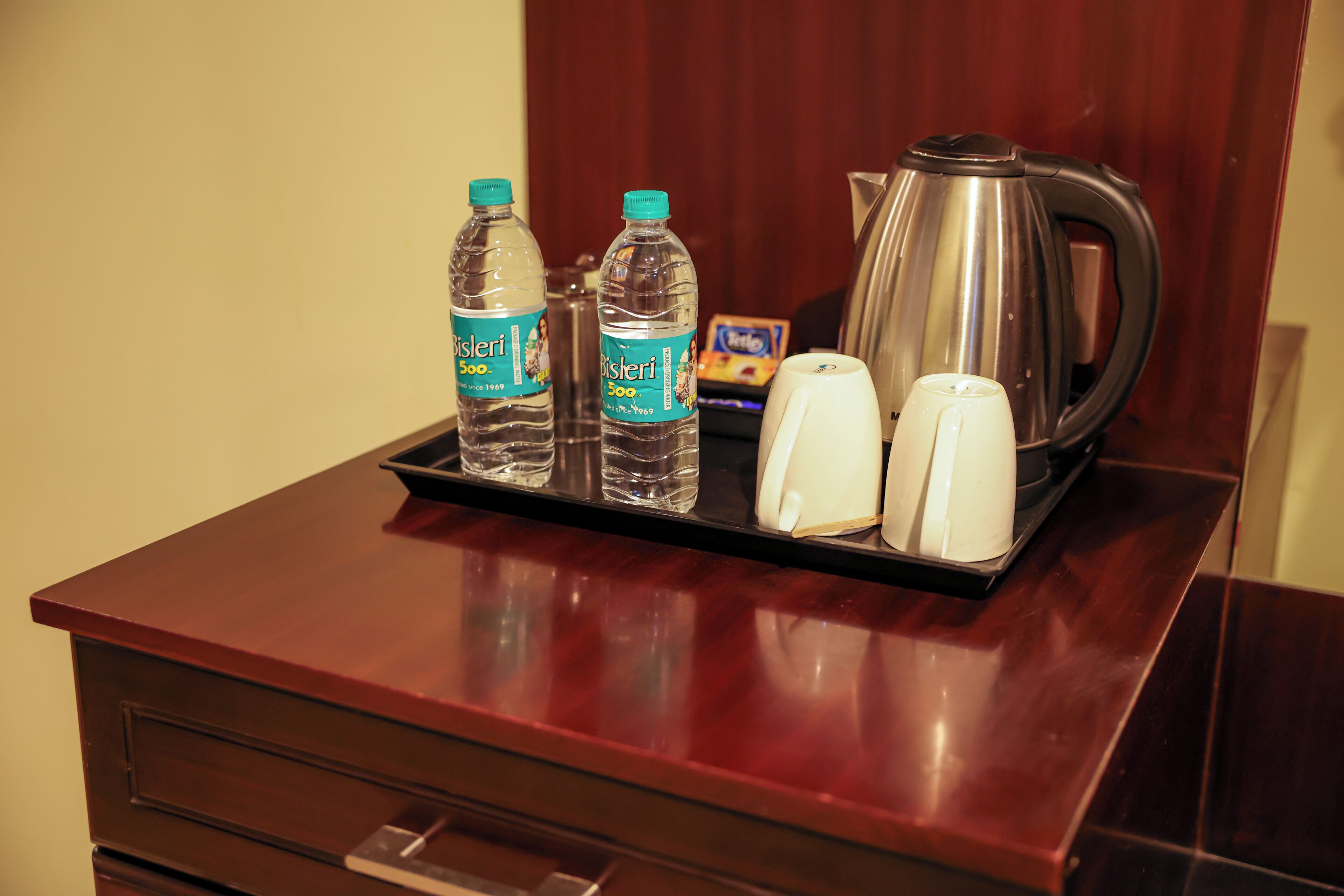Un plateau contenant deux bouteilles d’eau Bisleri, une tasse blanche et une bouilloire électrique argentée, posés sur une table en bois.