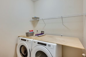 A laundry area with two front-loading Whirlpool washers, a wooden shelf, a metal drying rack, and a basket with laundry supplies. - Orange Park 'The Gathering House' w/ Pool Access (Orange Park)