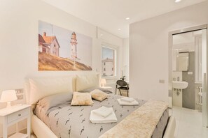 A bedroom with a bed, pillows, a nightstand, a painting, a chair, and a bathroom with a sink. - [ViaVeneto333]ModernApartment-FreeParking (La Spezia)