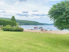 Une vue sur le lac, avec une zone herbeuse, un quai avec des bateaux et une colline boisée en arrière-plan.