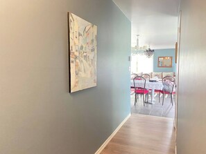 Un couloir mène à une salle à manger où se trouvent une table et des chaises, un lustre et un tableau accroché au mur.