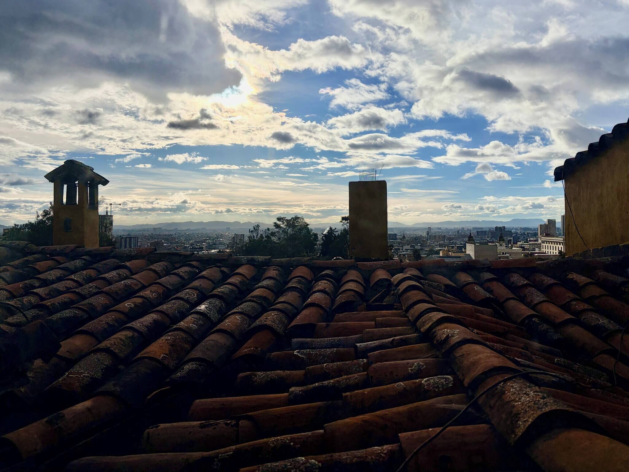 A rooftop with a terracotta tile roof, two small structures, and a cityscape in the background.