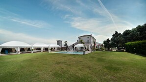 A grassy outdoor area with a pool, white tents, and a building with a tower.