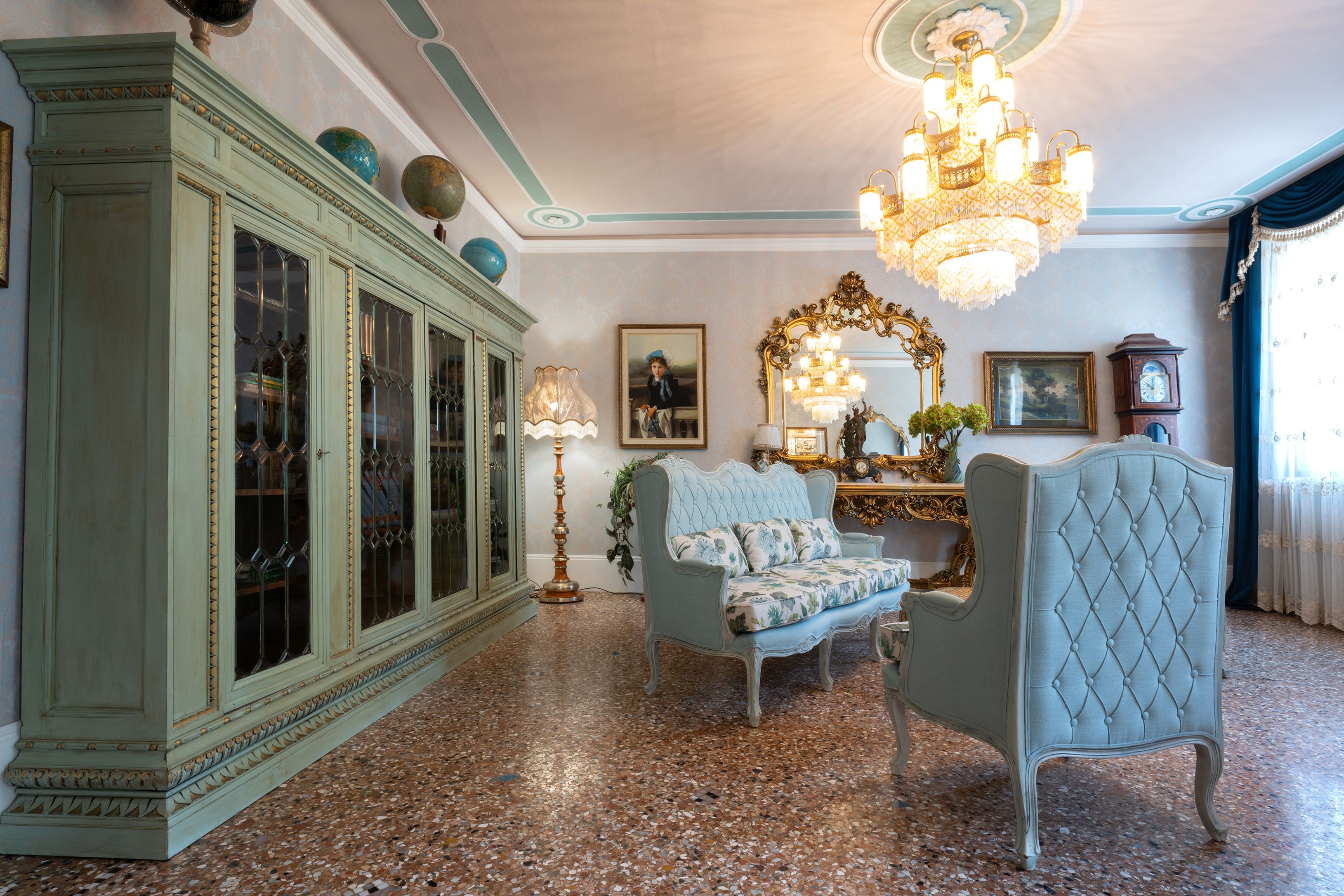 A room with a large green cabinet, a blue sofa, a decorative chair, a chandelier, and a clock on the wall.