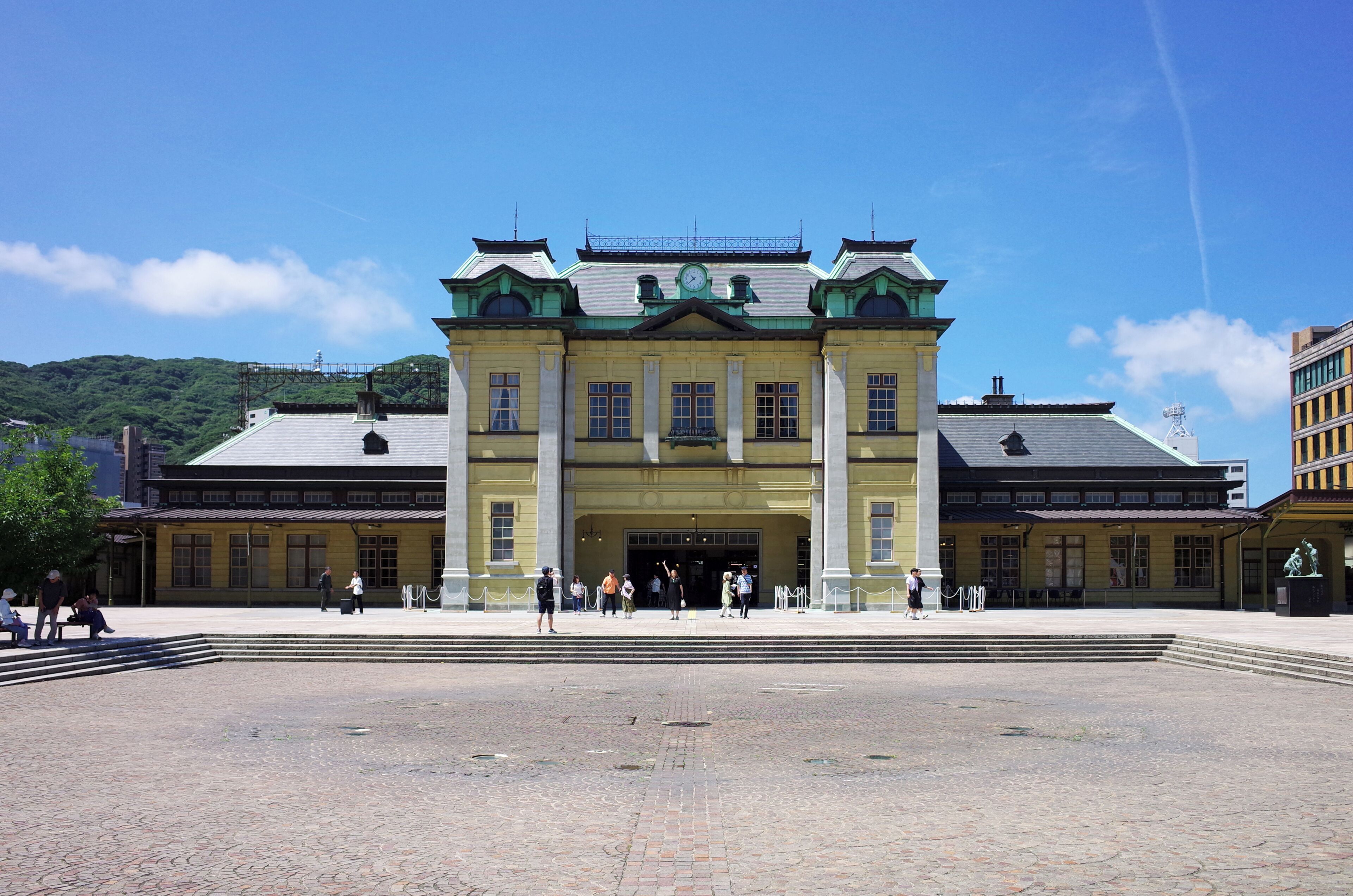 A historic building with a central entrance, flanked by two wings, and a paved plaza in front.