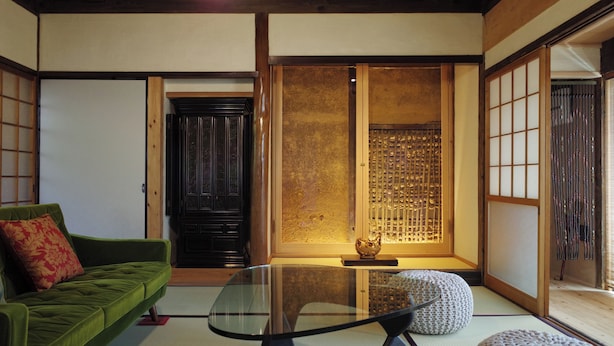 Um interior japonês tradicional com tatames, um sofá verde com uma almofada vermelha, uma mesa de vidro e um futum com um painel decorativo.