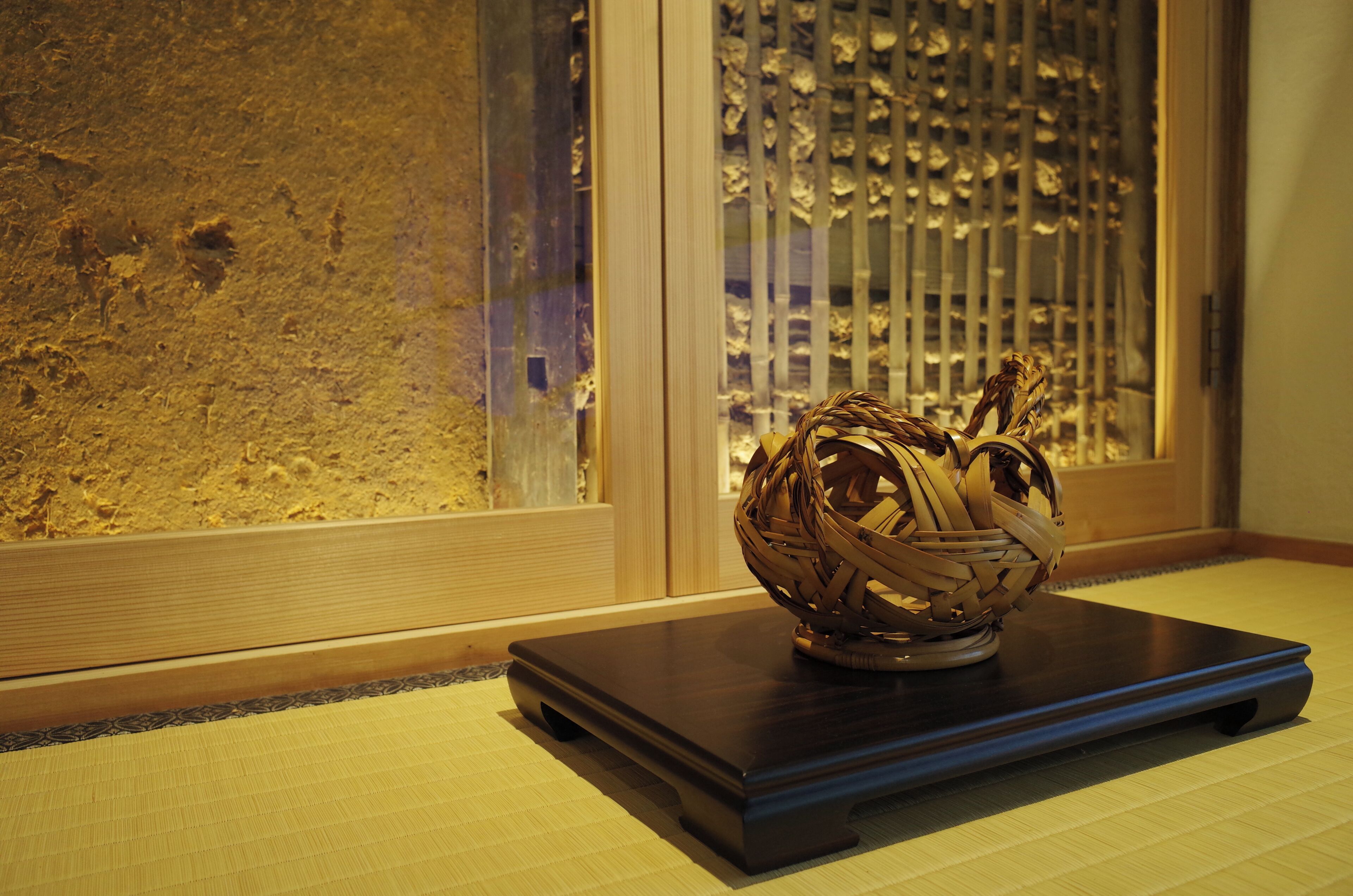 A traditional woven basket on a dark wooden stand in a room with tatami flooring and a shoji screen in the background.