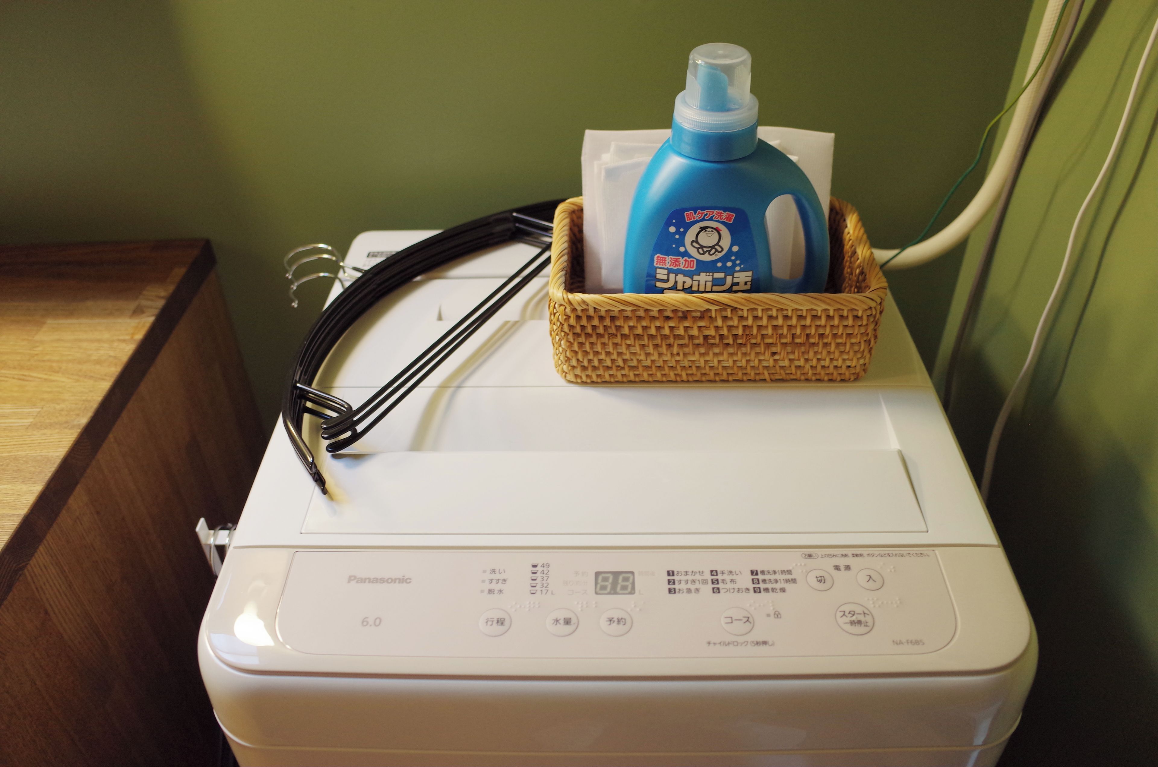 A front-loading washing machine with a control panel, a power cord, and a blue detergent bottle in a basket on top.