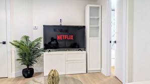 A modern living room with a flat-screen TV displaying the Netflix logo, a potted plant, a glass coffee table, and a white entertainment unit.