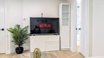 Un salon moderne avec un téléviseur à écran plat affichant le logo de Netflix, une plante en pot, une table basse en verre et un meuble de rangement blanc pour appareils multimédias.