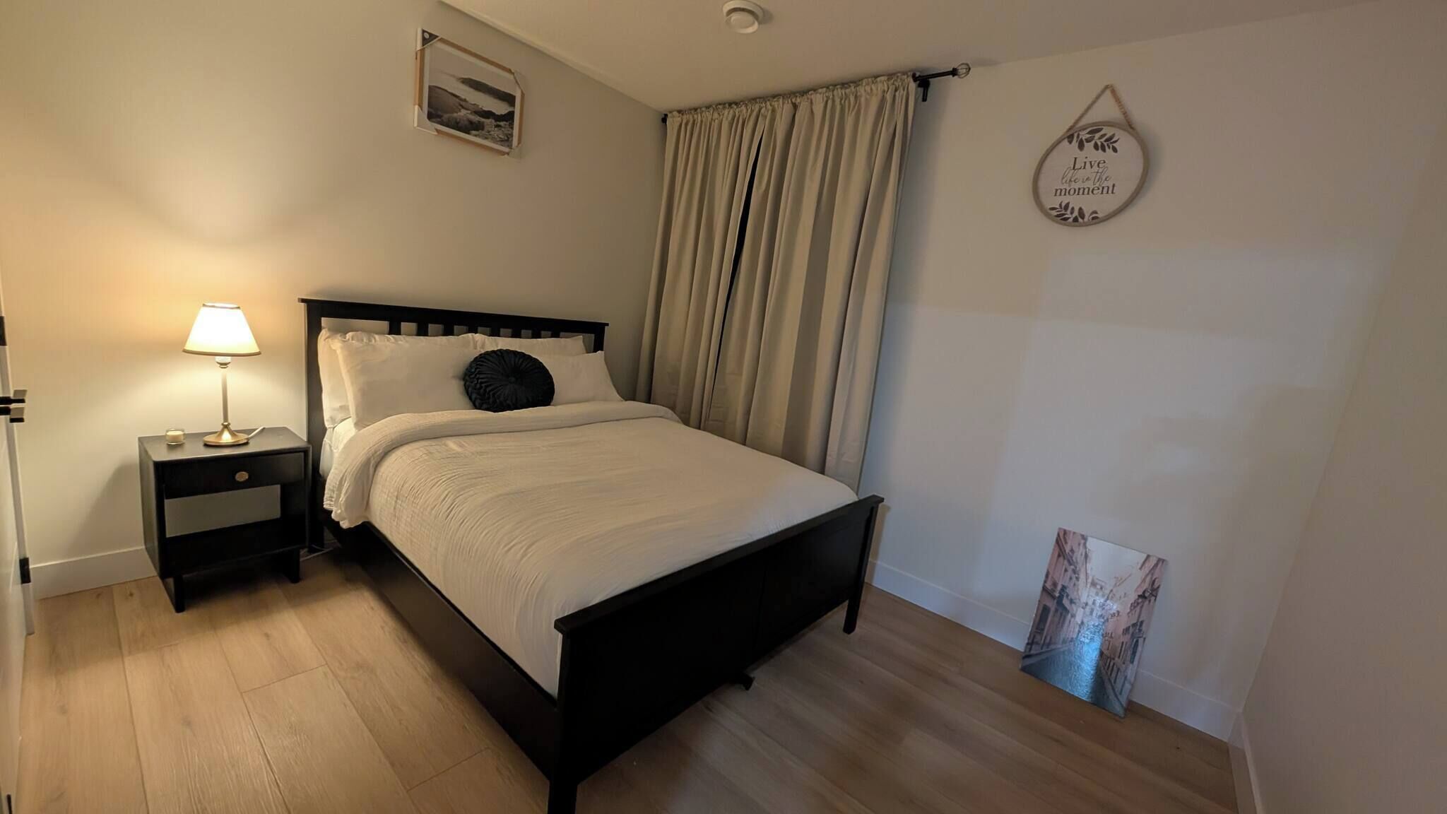 Una cama bien hecha con cabecera, una mesita de noche con lámpara, y una pared con un cuadro enmarcado y una frase motivacional.