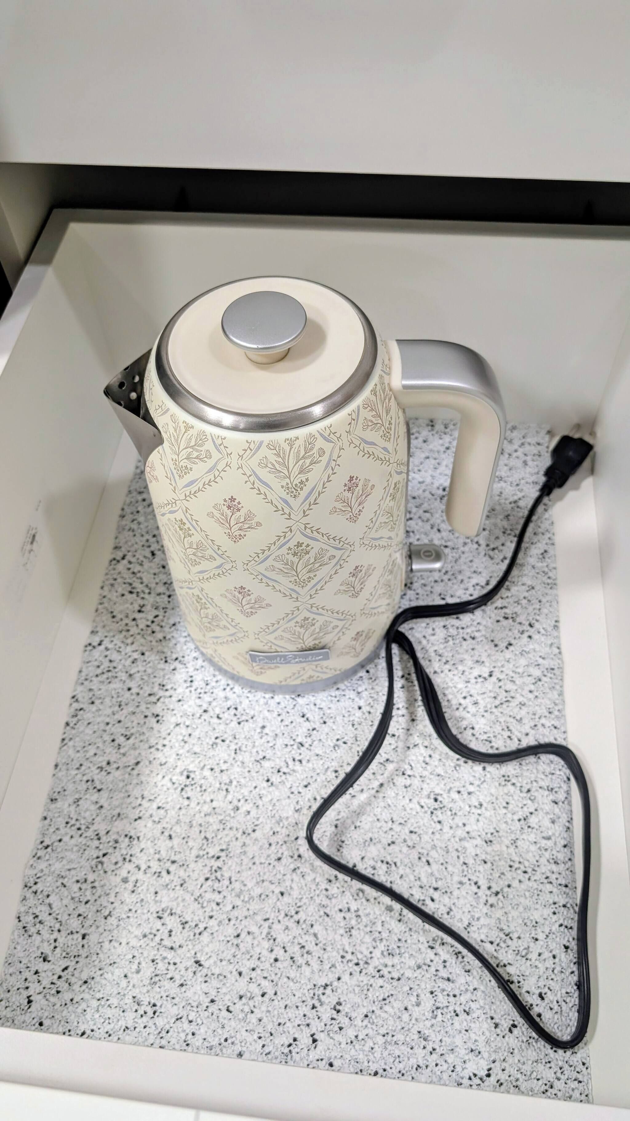 Una tetera blanca con motivos florales y un cable de alimentación conectado, sobre una encimera con motas.