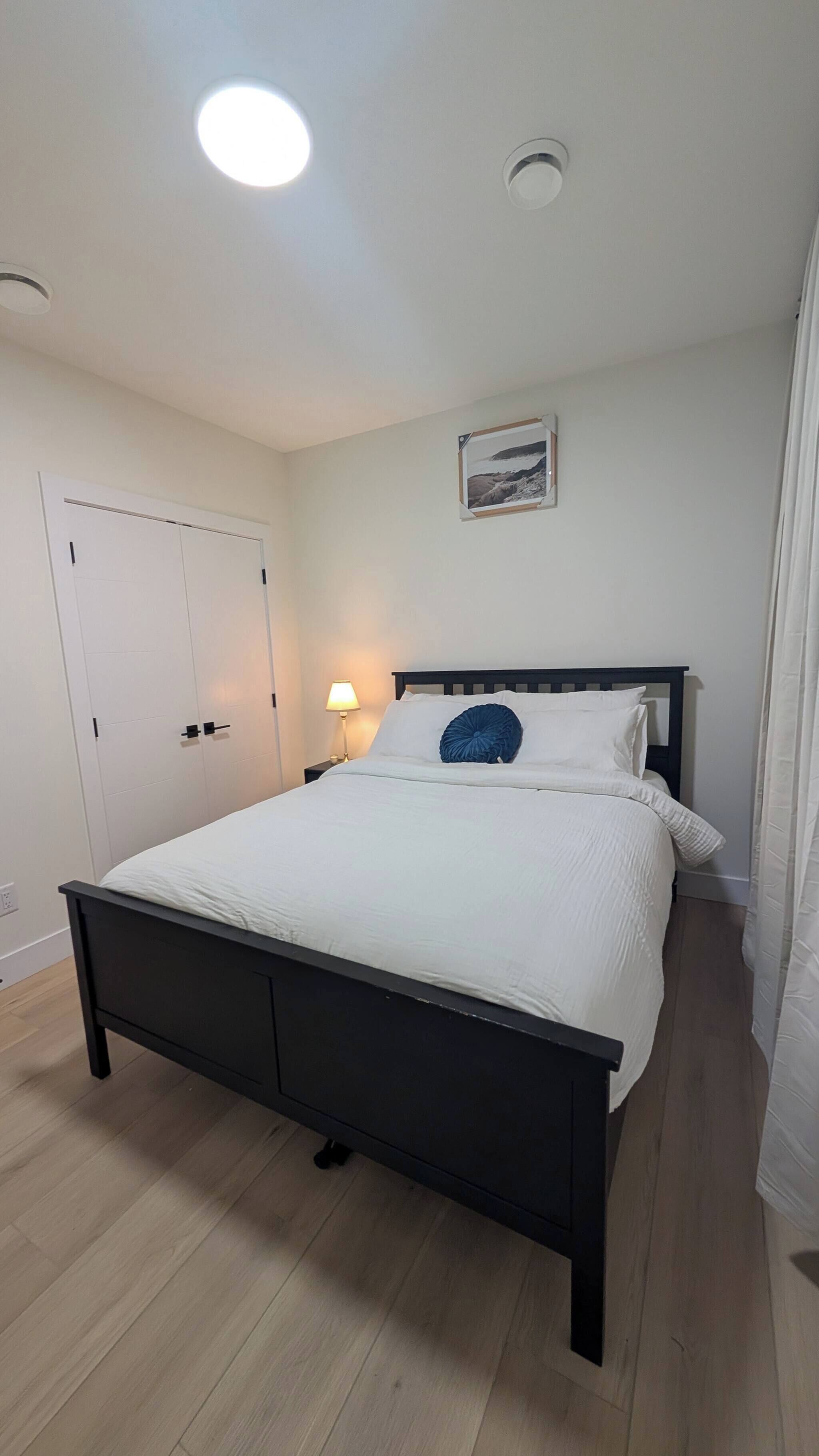A bedroom with a bed, a nightstand, a lamp, a door, and a ceiling light.
