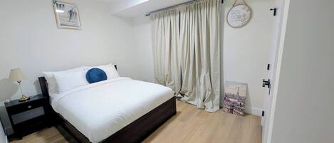 A bedroom with a bed, a nightstand, a lamp, a window with curtains, and a door.