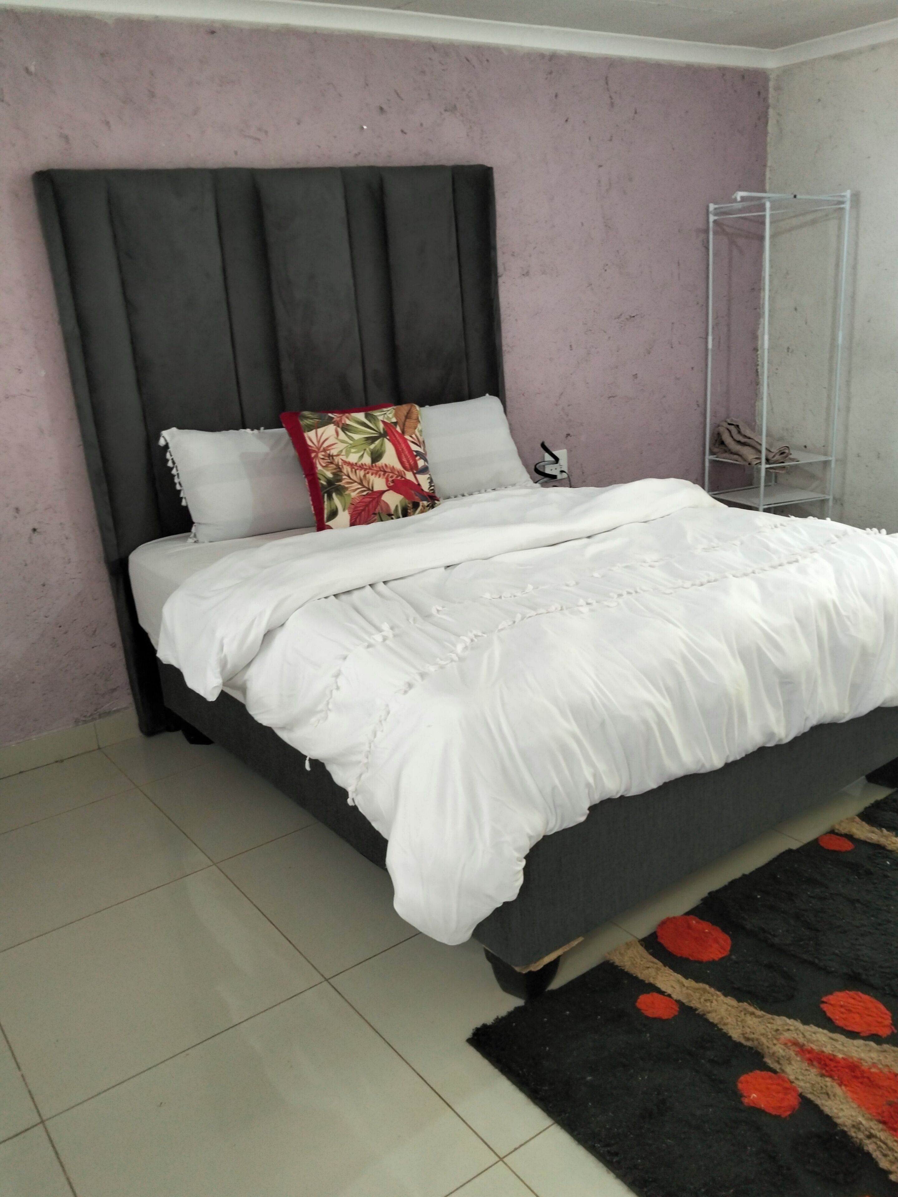 A bed with a grey headboard, white bedding, and a black and red patterned rug.