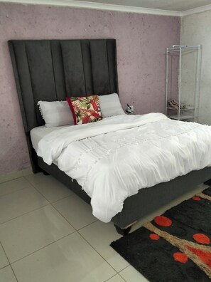 A bed with a grey headboard, white bedding, and a black and red patterned rug.