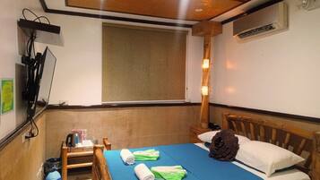 A hotel room with a bed, a wooden headboard, a window with a blind, a wall-mounted TV, and a small table with a kettle and a trash bin.